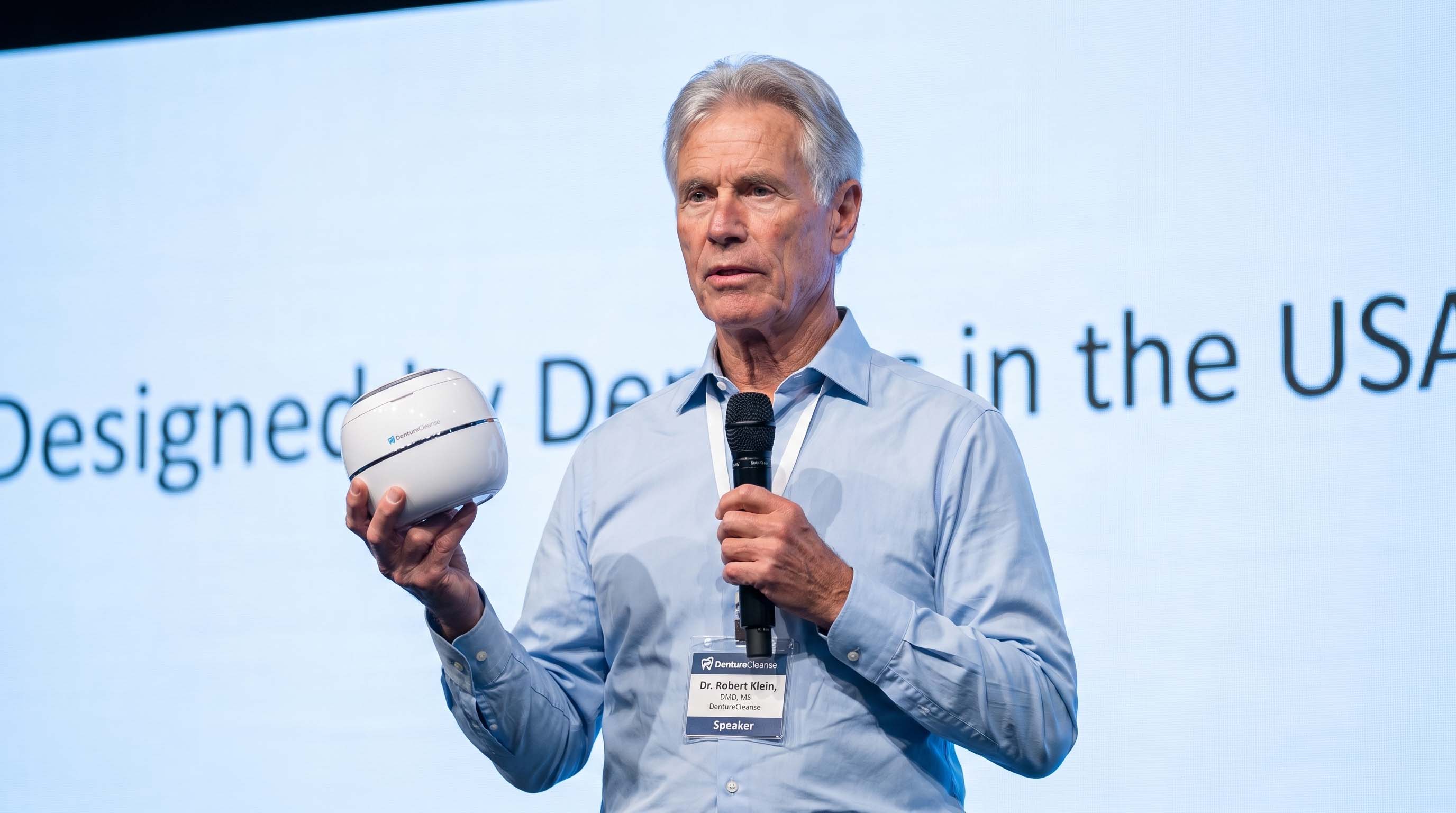 Dr. Robert Klein, DMD, MS, presenting the DentureCleanse pod on stage beneath a screen reading Designed by Dentists in the USA